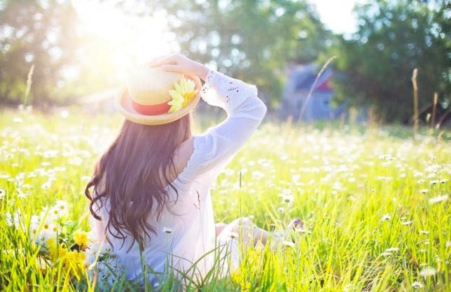 Frau mit weissem Hemd und Sonnenhut sitzt auf hoher Wiese und blickt in die Sonne