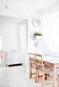 Eine offene, helle Küche mit zwei Stühlen, die vor einem Tisch stehen, grüner Pflanze an der Wand, einer weißen Uhr und im nächsten Raum eine weiße Kommode beachtet alle Regeln der gesunden Raumgestaltung.