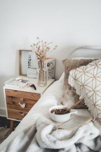 Ein Bett, Nachtkästchen mit Trockenpflanzen, einem Bild und Büchern darauf sind in Naturfarben gehalten und Teil einer gesunden Raumgestaltung.