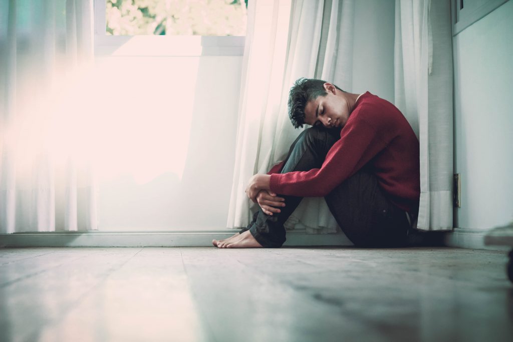Junger Mann sitzt in Wohnung auf dem Boden und umarmt seine Beine weil er unter Angststörungen leidet