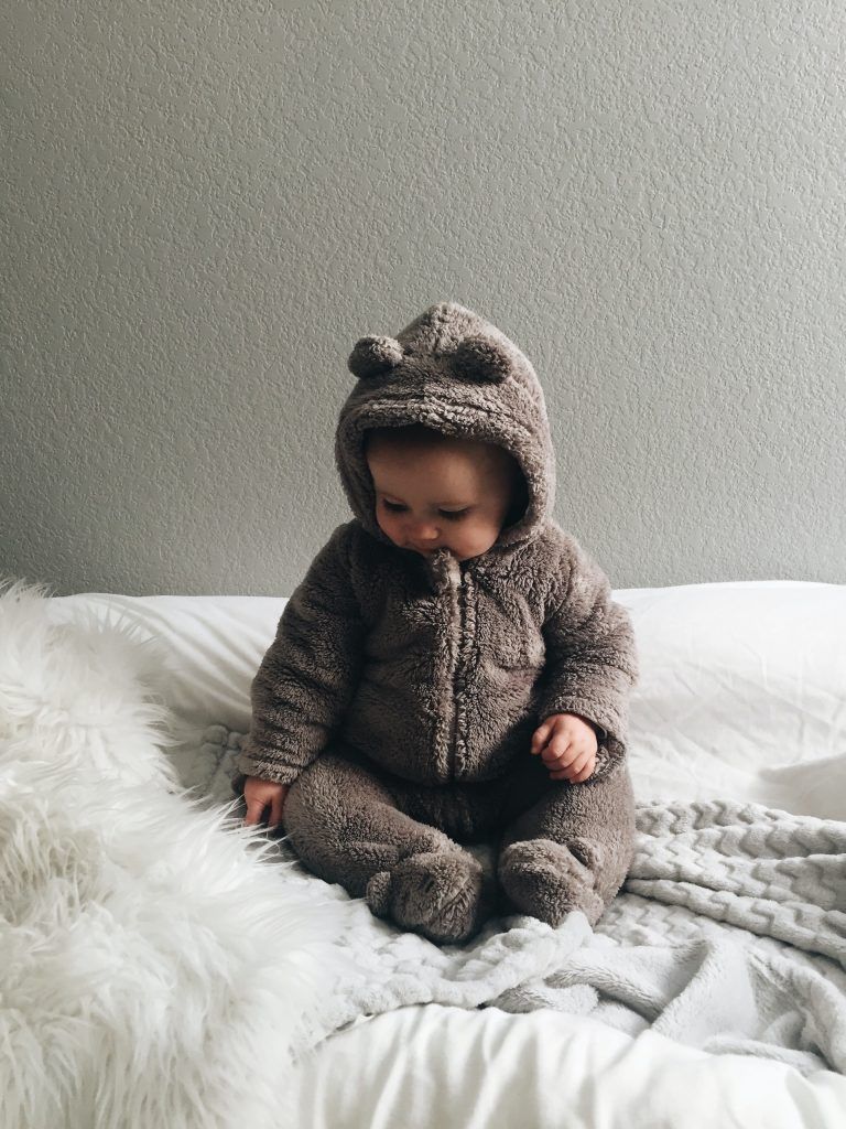 Ein Baby mit braunem Bären-Overall sitzt auf einem weißen Bett vor einer grauen Wand.