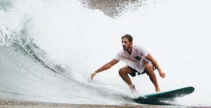 Abnehmen mit Sport: Ein Surfer im weißen Hemd, der gerade auf einer hohen Welle vor einer Klippe surft, kümmert sich um die richtige Sporternährung.