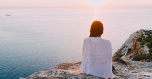 Frau sitzt auf Felsen und blickt auf Sonnenaufgang über dem Meer, während sie über die Psychologie des Abnehmens nachdenkt.